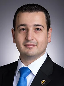 Man in a suit and blue tie, facing forward, with short dark hair, on a gray gradient background.