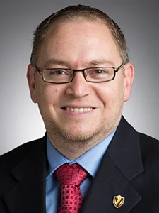 Smiling man in a suit with glasses and a red tie, against a plain gray background.