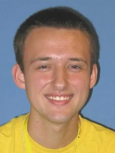 Young man with short brown hair smiles, wearing a yellow shirt, in front of a blue background.