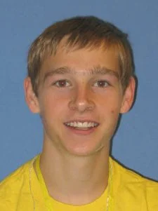 Smiling teenage boy with short light brown hair, wearing a yellow shirt, against a blue background.