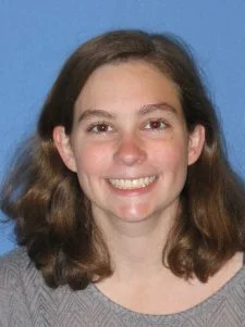 A woman with brown hair smiles at the camera against a blue background.