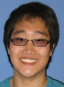 A person with short dark hair and glasses smiling in front of a blue background.