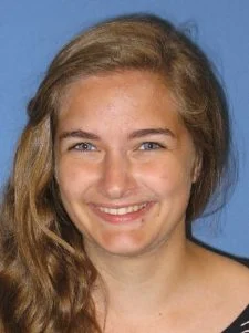 A young woman with long, wavy light brown hair smiles in front of a blue background.