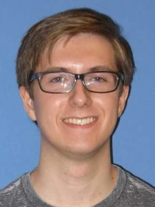 Young person with short brown hair, glasses, and a gray shirt smiling in front of a blue background.