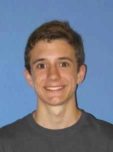 Young man with short brown hair, smiling, wearing a dark shirt, in front of a solid blue background.