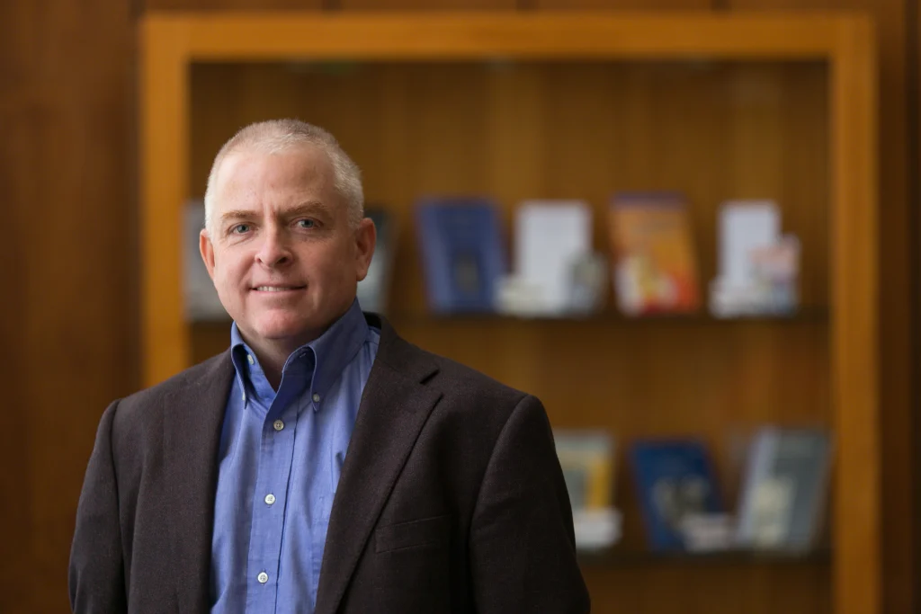 Man in a blue shirt and dark blazer stands smiling in front of a bookshelf with blurred books, featured in CC Faculty News.