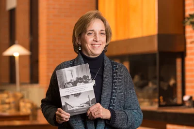 Woman smiling and holding a book while standing indoors in a warmly lit room, featured in CC Faculty News.