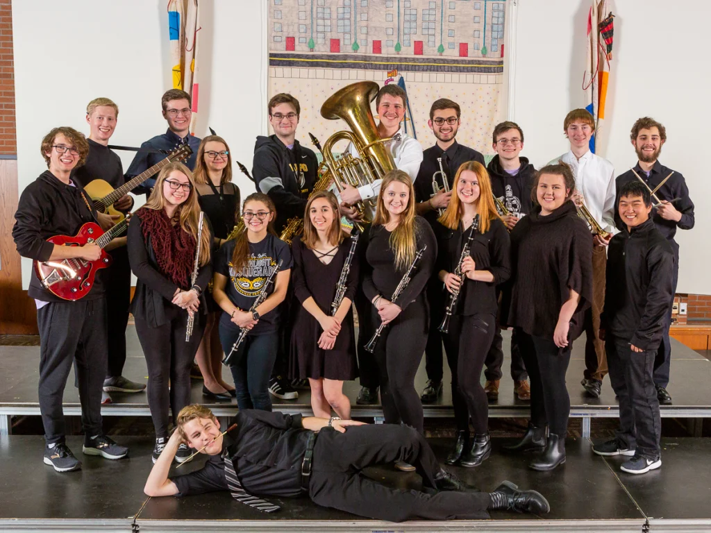 A group of students with musical instruments pose together on stage, smiling at the camera during their First-Year Production.