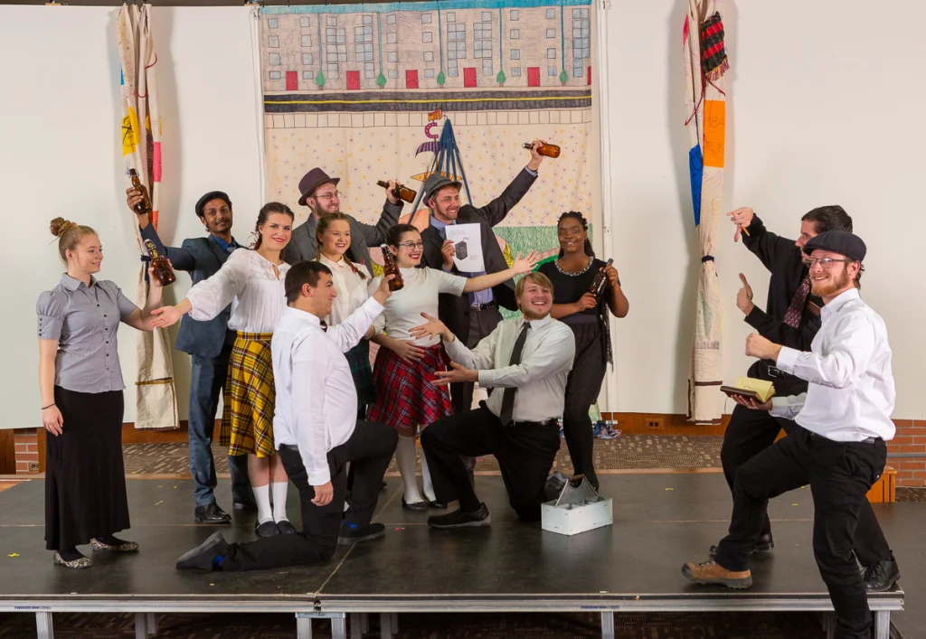 A group of people in costumes pose cheerfully on a stage during the First-Year Production, some holding bottles, in front of a colorful backdrop.