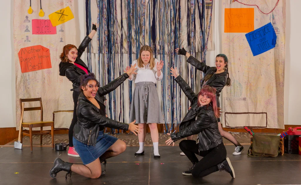 Five women pose on stage in this First-Year Production; one in the center looks sad, while four around her gesture dramatically and smile.
