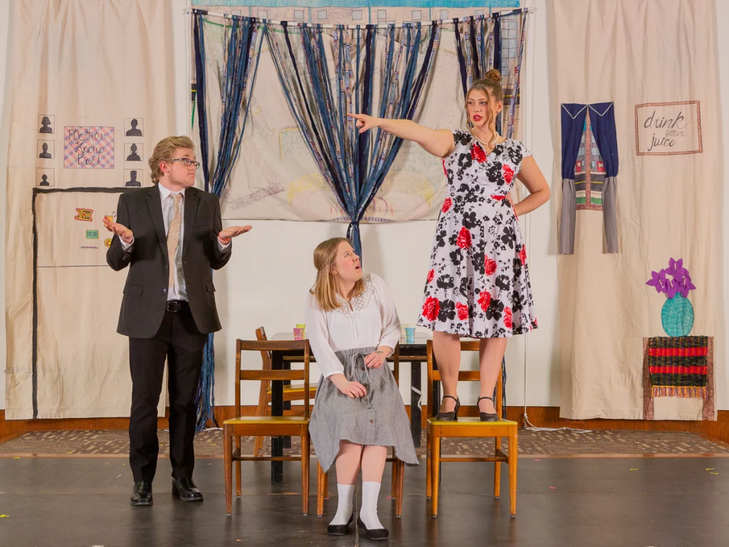 In this First-Year Production, three people act on stage; one woman in a floral dress stands on a chair, pointing and speaking.