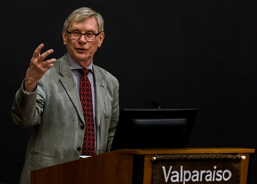 Older man in a gray suit speaking at a podium with "Valparaiso" on the front, gesturing with one hand, during a 2018 event.