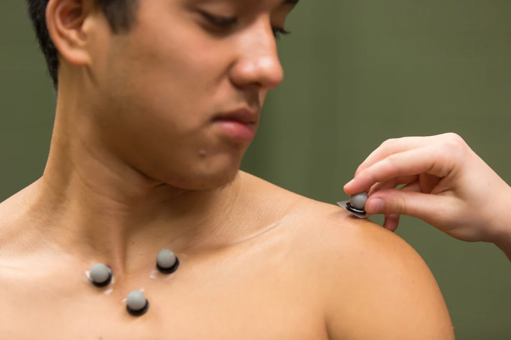 A person places sensor electrodes on a man's chest and shoulder for a medical or physical test, showcasing a student opportunity to gain hands-on experience in an innovative degree program.