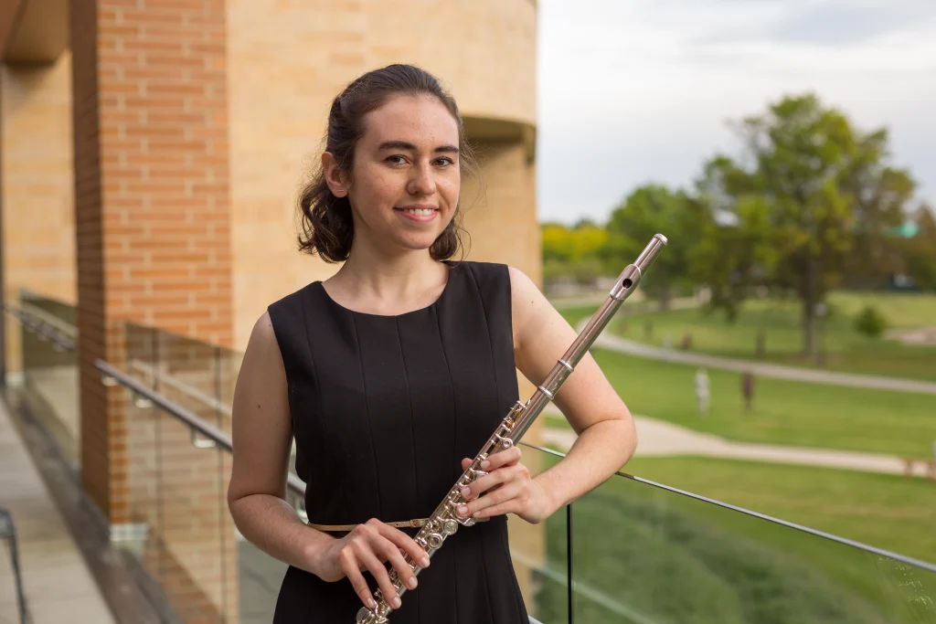 Young woman in a black dress holding a flute, standing on a balcony with a grassy park in the background, showcasing her passion for music.