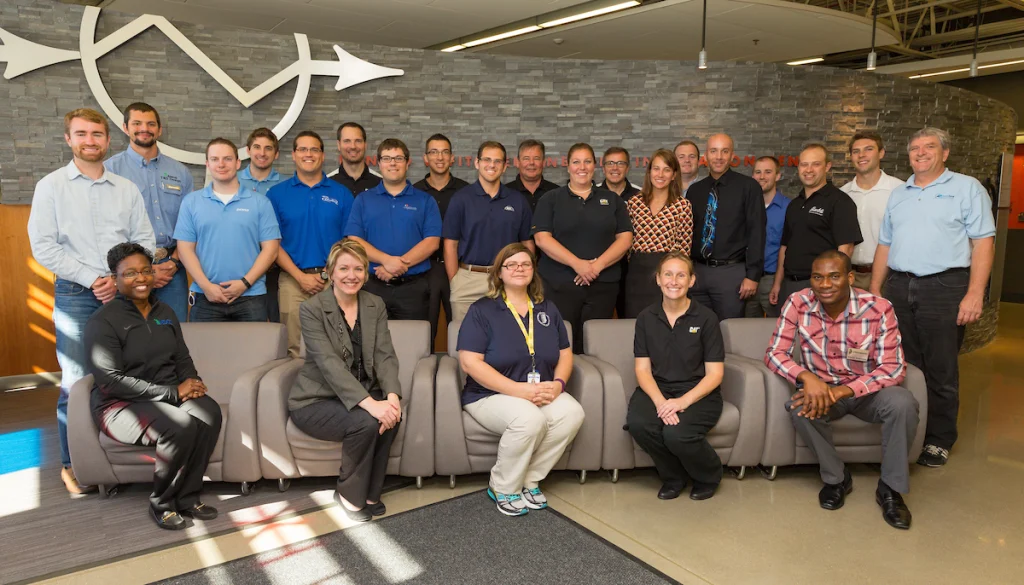 A group of twenty-four adults, some seated and some standing, pose together in a modern office lobby during a College of Engineering Career Fair.