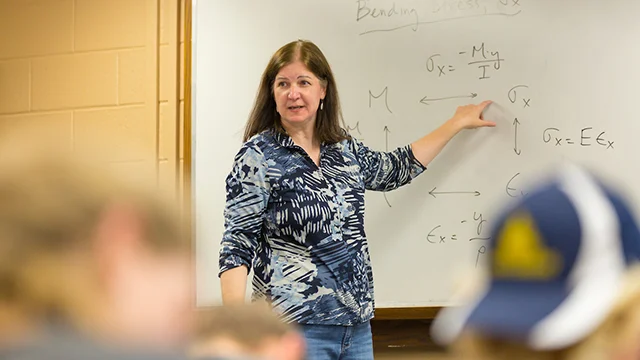 A woman, the new chair of the Civil Engineering department, points to equations on a whiteboard while teaching a classroom of students.