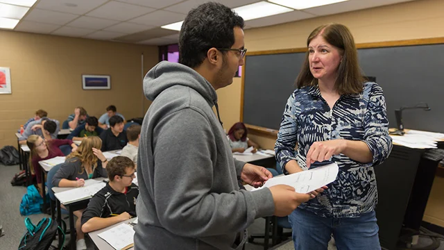 A teacher, possibly the Department Chair of Civil Engineering, hands papers to a student at the front of a classroom while other students work at their desks.