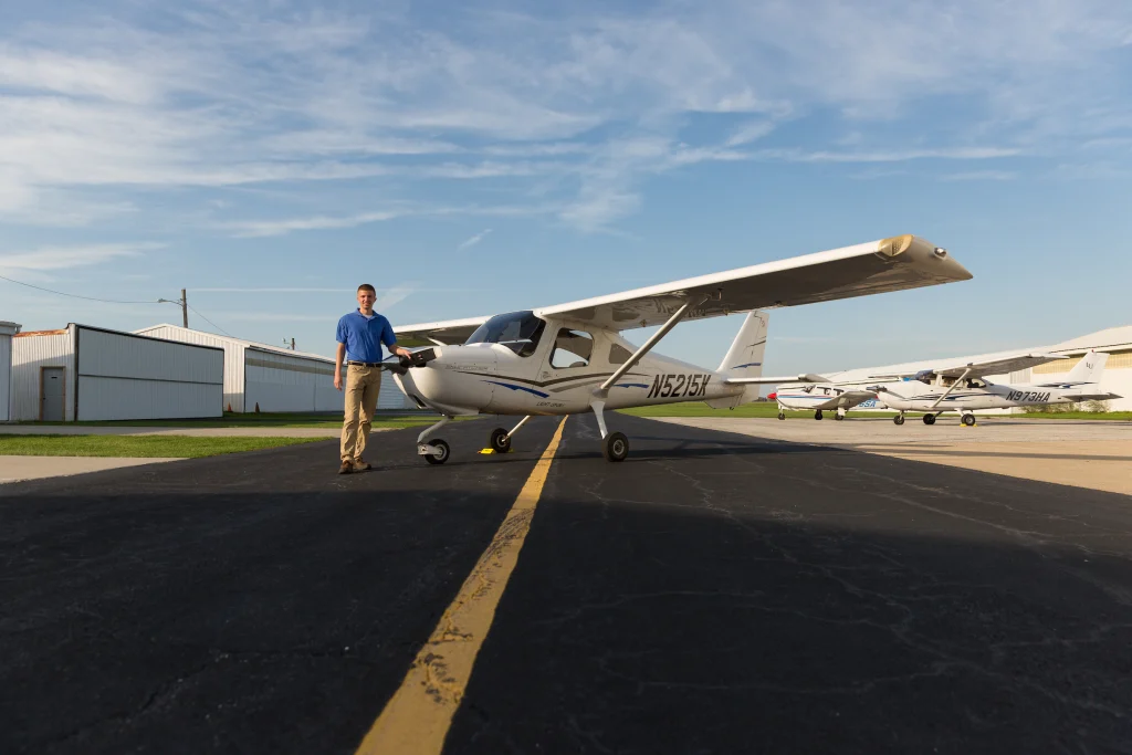 A person stands next to a small white airplane on an airport tarmac under a blue sky, ready to launch their journey with the Aviation Program debuting Fall 2017.