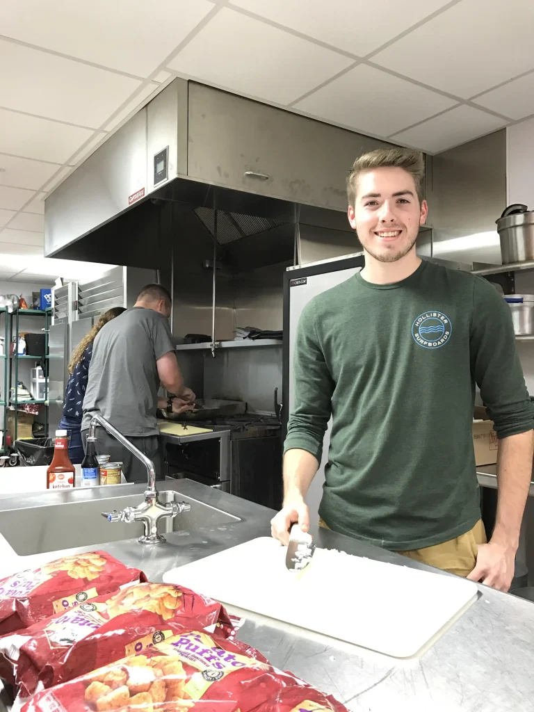 Young man smiling and chopping vegetables in a commercial kitchen with two people cooking in the background.