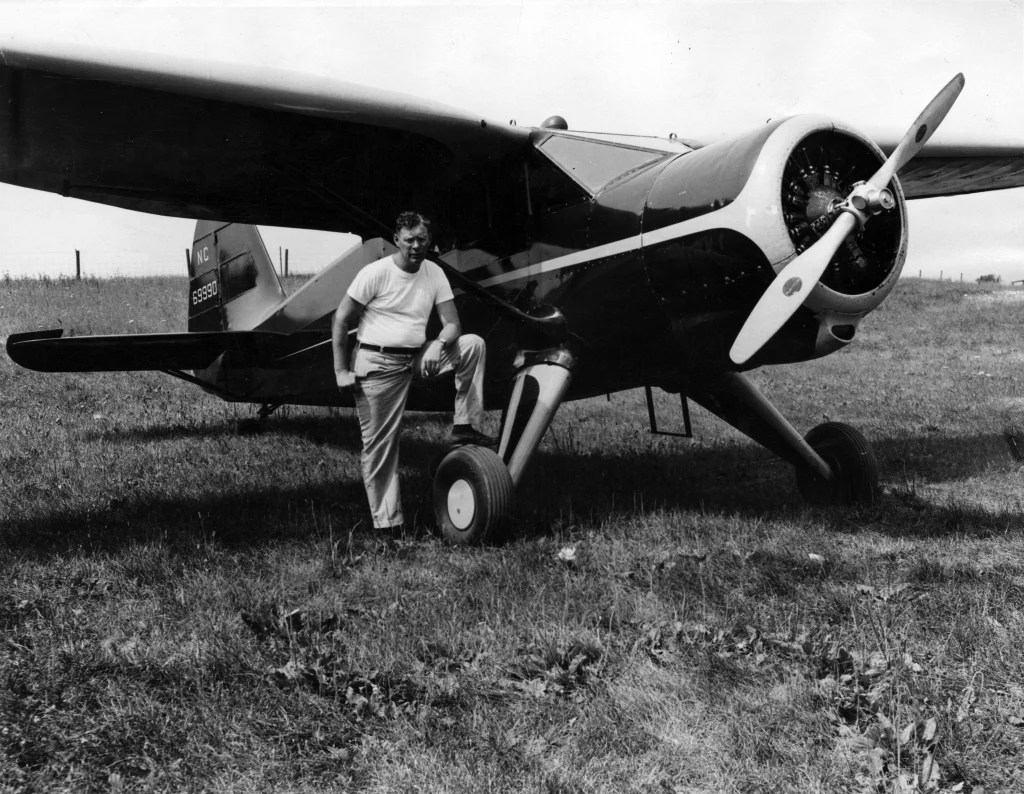 Man standing with one foot on the wheel of a small propeller airplane parked on a grassy field, ready to launch his aviation journey as part of the Fall 2017 Aviation Program.