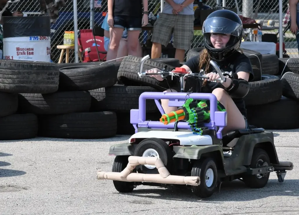 Girl wearing a helmet drives a toy car with a water gun mounted on front, tires stacked in background.