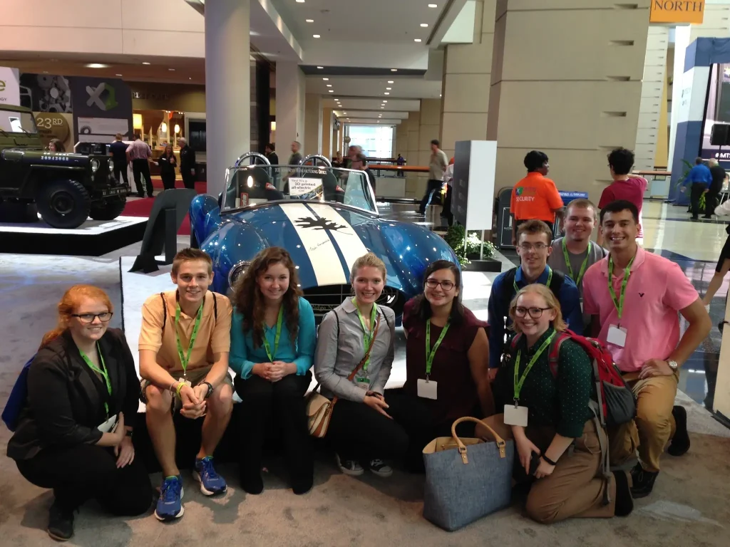 A group of smiling young adults pose in front of a blue race car at an indoor event or expo.