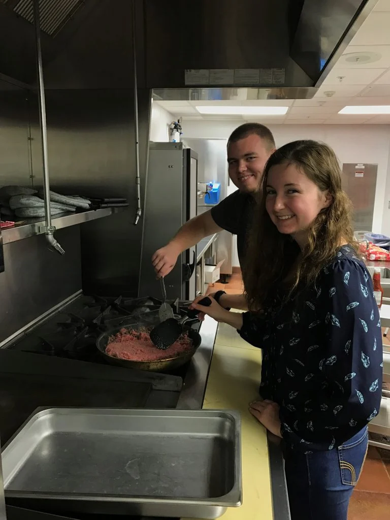 Two people cooking ground beef in a pan on a stove in a commercial kitchen, both smiling at the camera during Dinner with the Professors.