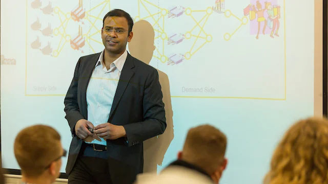 A College of Business professor in a suit presents to an audience in front of a screen displaying a network diagram, highlighting insights from this crucial field.