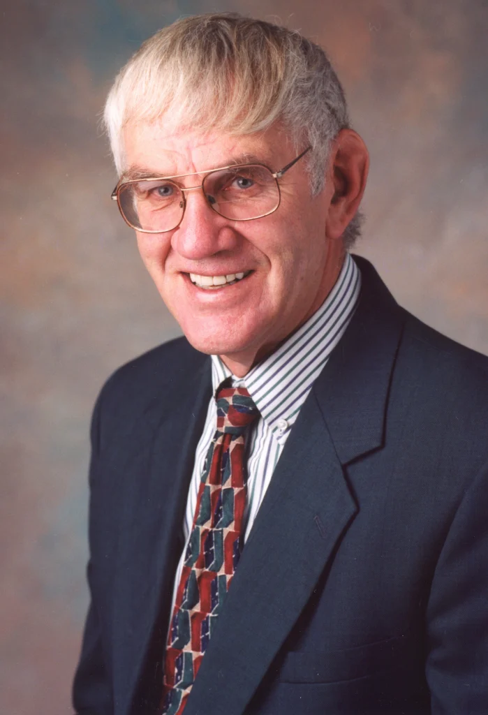 An older man, identified as Arlin Glenn Meyer, wearing glasses, a suit, and a patterned tie, smiles at the camera against a studio background—capturing a warm moment for his memorial.