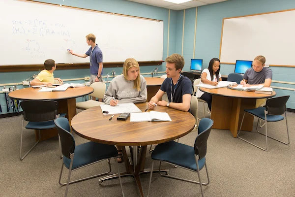 Students working in groups at round tables in a classroom at the Hesse Center, with one student writing on a whiteboard as part of the anniversary celebration.