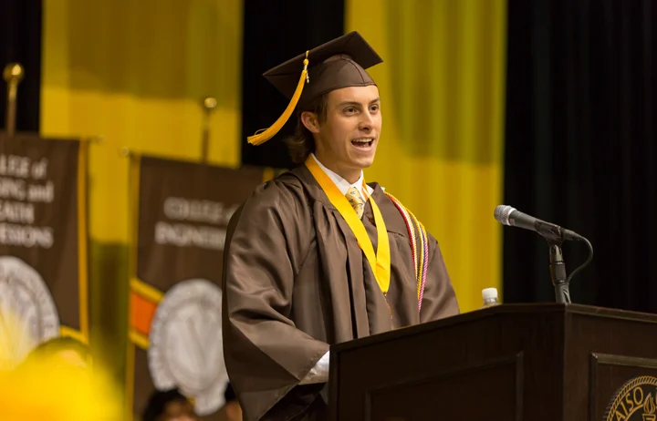 A 2017 graduate in cap and gown shares their future graduate plans while speaking at a podium during a graduation ceremony.