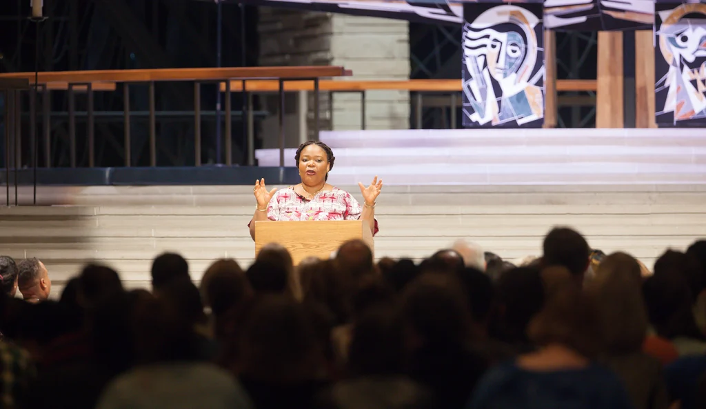 A woman speaks at a podium to an audience in a campus auditorium or church, with abstract art behind her—addressing the crowd as a distinguished Nobel Peace Laureate.