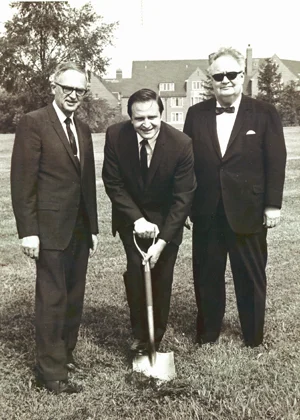 Three men in suits pose on grass at Christ College; the man in the center holds a shovel, marking new beginnings, as if for a groundbreaking ceremony.
