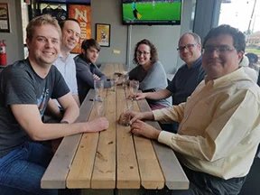 Six people smiling, sitting around a wooden table in a casual restaurant with empty glasses on the table—members of CC Alumni Reading Groups reconnecting and sharing stories from 2001-2019.