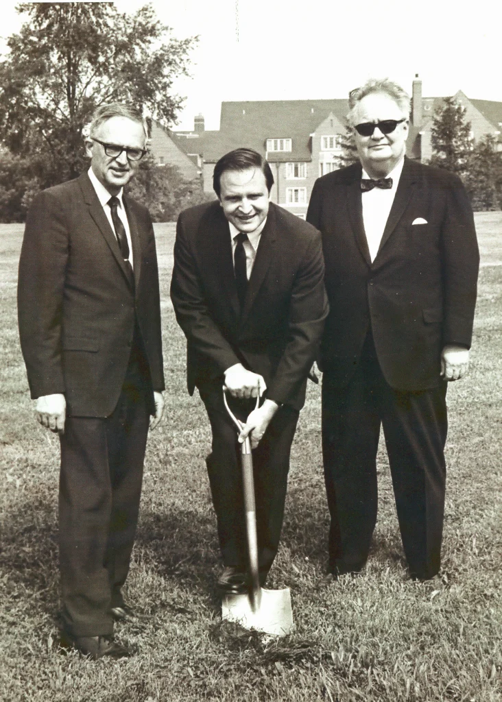 Three men in suits at a groundbreaking ceremony for CC The Beginning Years; one is holding a shovel in a grassy field.