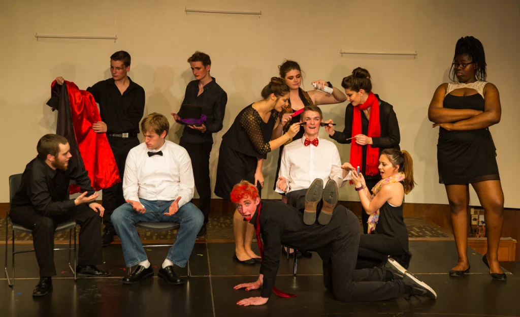 A theater group, much like a college orientation, prepares backstage—dressing and applying makeup to two seated men in white shirts and bow ties—capturing the excitement of a student transition into new experiences.