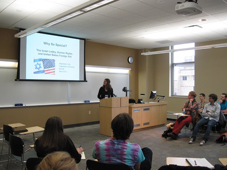 A woman presents research about Israeli and U.S. foreign aid to a small group in a classroom, sharing her findings as part of an NCUR-style lecture.
