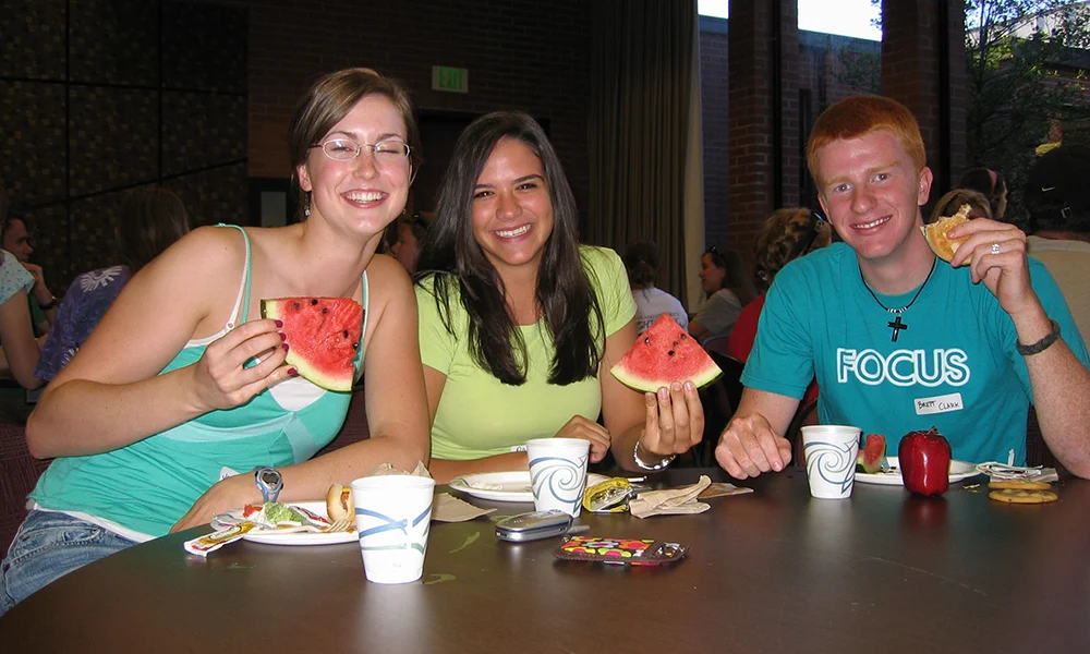 Three smiling young people sit at a table eating watermelon and snacks in a casual indoor setting, capturing the fun and connections that define Student Life, both past and present.