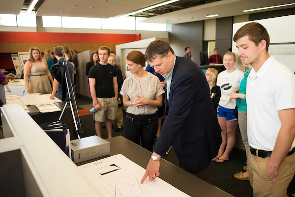 A group of people gathers around a man pointing at papers on a table in a modern office space, discussing Senior Design Capstone Projects, live captured during Spring 2017.