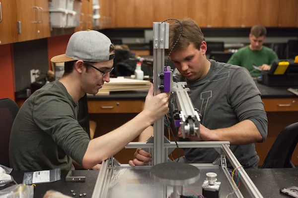Two young men, embodying Global Engineers, assemble a metal device in a science lab, drawing on their international experience as others work diligently in the background.
