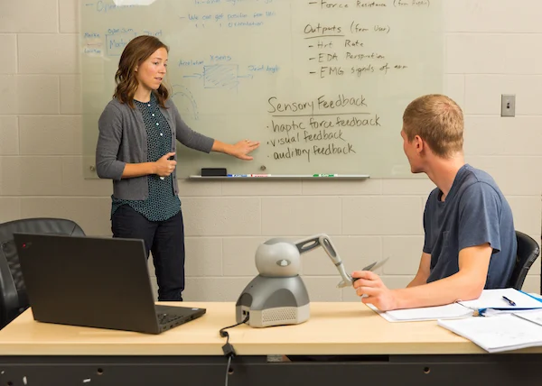 Professor Reva Johnson discusses bioengineering research as she points to notes on a whiteboard, while a man listens; a laptop and device sit on the table.
