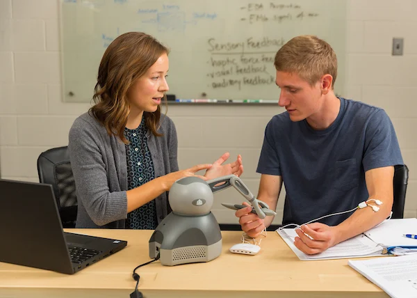 Two people use a medical simulation device at a desk with a laptop and papers, engaging in cutting-edge research in bioengineering within a classroom setting.