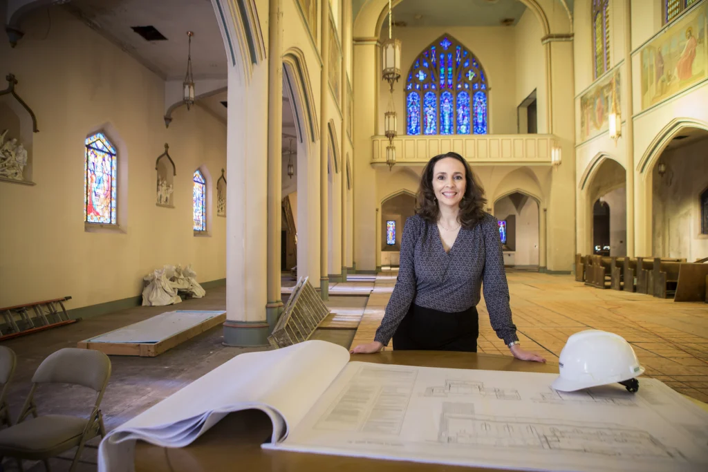 A Valpo Alum stands by architectural plans in a large, ornate church under renovation, with stained glass windows behind her, helping to build a world of difference on a solid foundation.