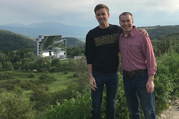 Two men smiling in front of a green landscape with a large mirrored building, showcasing the spirit of Global Engineers and their international experience.