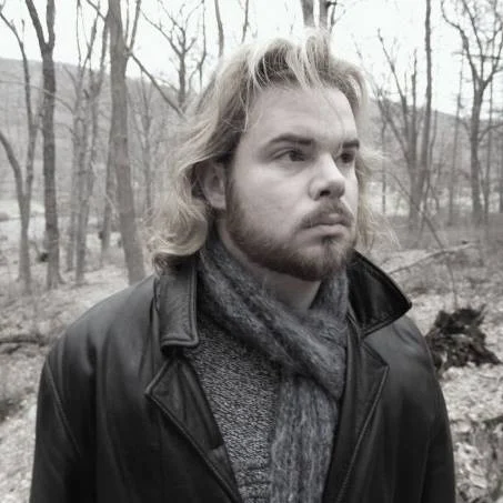 A man with a beard and long hair, wrapped in a scarf and leather jacket, stands in a leafless forest—an image evoking the introspective mood of poetry from the pages of Valparaiso Poetry Review.