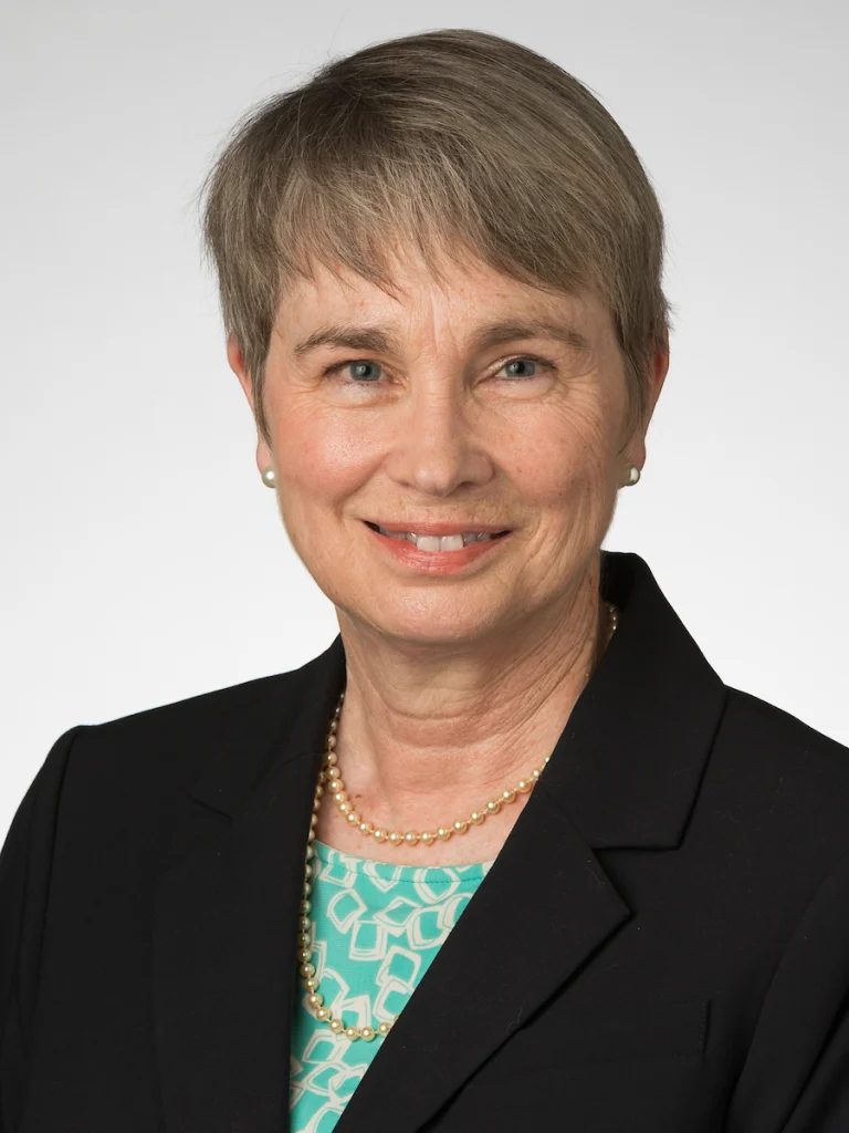 Smiling older woman with short gray hair in a black blazer and patterned teal top, against a light background, representing the distinguished faculty of Valparaiso University’s Christ College – The Honors College.