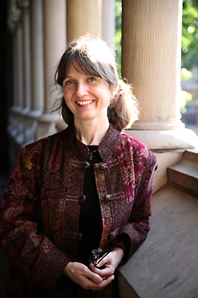 A woman in a patterned jacket, possibly at Valparaiso University, smiles while standing by columns as sunlight streams in behind her, reflecting the spirit of Religion and the Healing Arts.