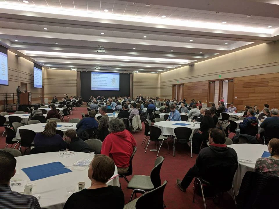 Large group of people seated at round tables in a conference room at Valpo, attending the Mathematical Association of America Trisection meeting and listening to a speaker at the front.