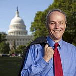 Smiling undergraduate in a blue shirt and red tie stands outside, holding his jacket, with the U.S. Capitol behind him, perhaps preparing for a commencement address or reflecting on the World Food Prize.