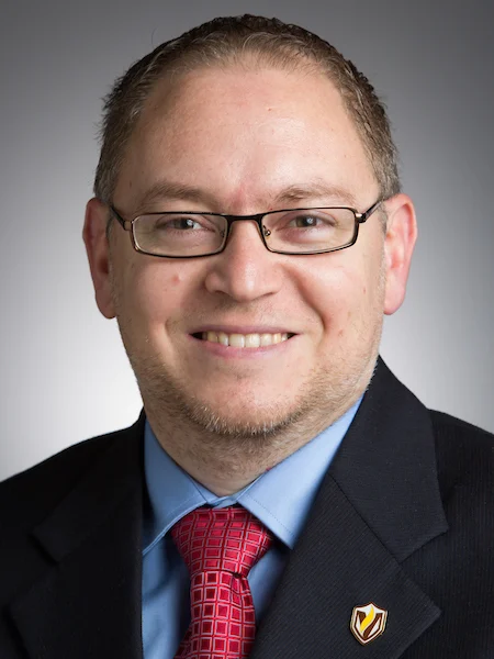 Smiling man in glasses, suit, and red tie, posed against a neutral gray background, representing the excellence in teaching shown by Valpo professors.
